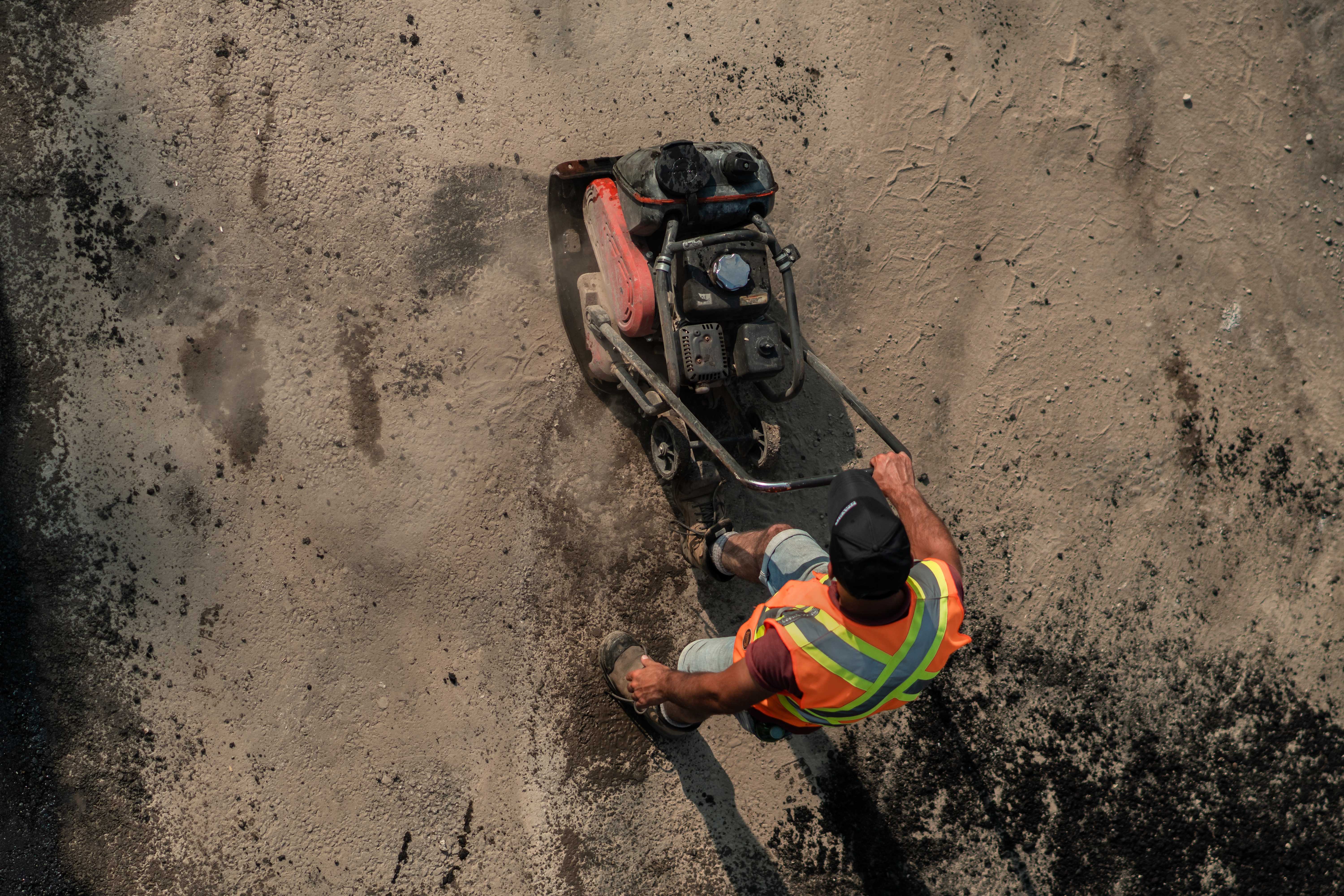 Overhead view of a contstruction worker sanding a rough road surface with a piece of equipment.