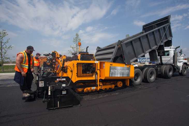 A rear-view large dump truck emptying loose asphalt into a piece of paving equipment as several construction workers work nearby.