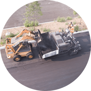 An aerial video of a piece of paving equipment scooping asphalt from the back section of another large vehicle.