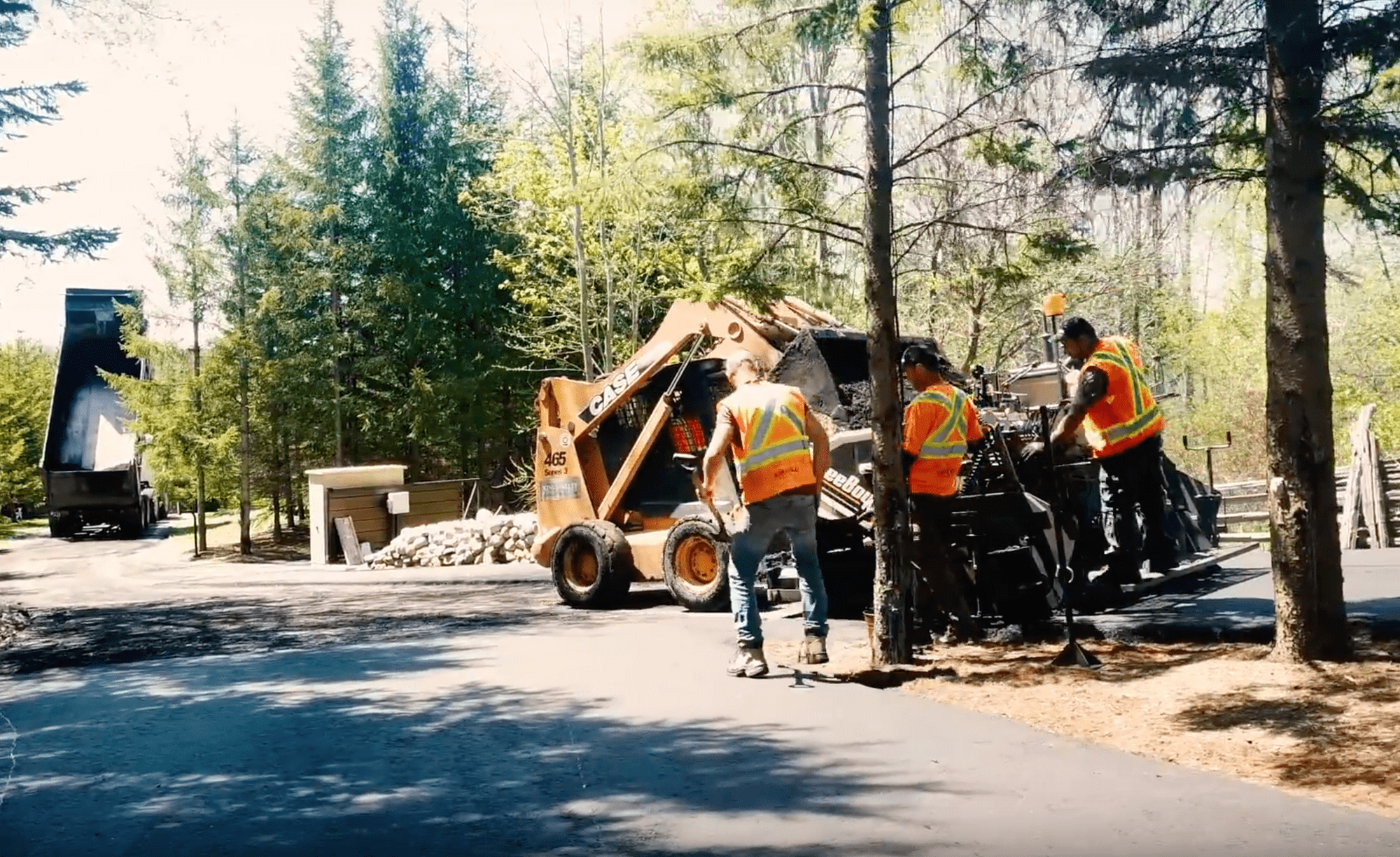 Three construction workers using large equipment to work on a large driveway on a sunny day.